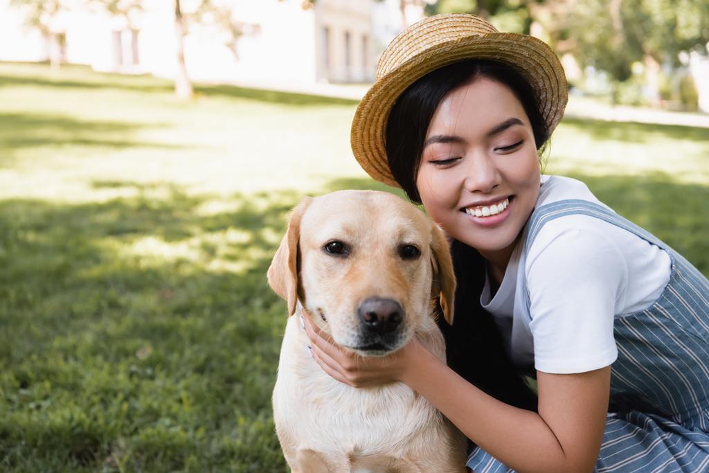 felice donna asiatica in cappello di paglia abbracciando labrador giallo nel parco - Foto, immagini