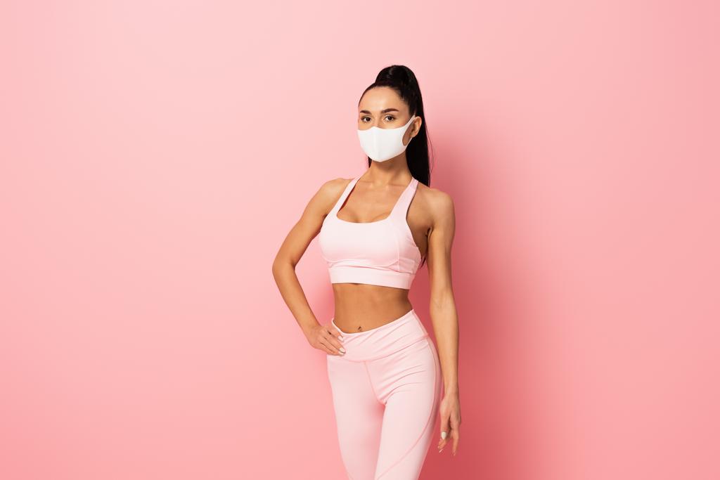 jonge sportvrouw in medisch masker staan met de hand op heup op roze  - Foto, afbeelding