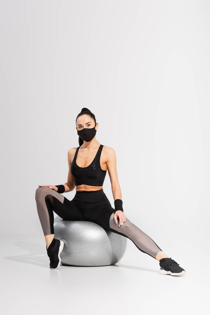 brunette sportvrouw in zwart beschermend masker oefenen op fitness bal op grijs - Foto, afbeelding