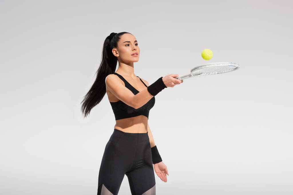 молодая спортсменка, держащая теннисную ракетку и мяч, играя изолированно на сером - Фото, изображение