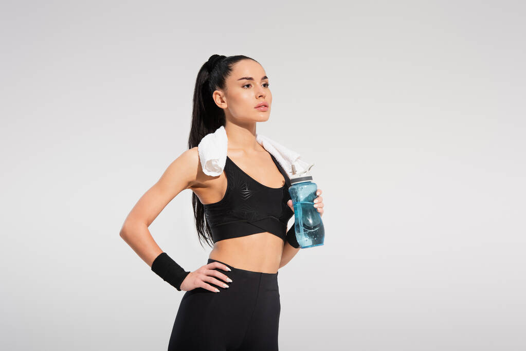 young sportswoman with towel on shoulders holding sports bottle while posing with hand on hip isolated on grey - Photo, Image