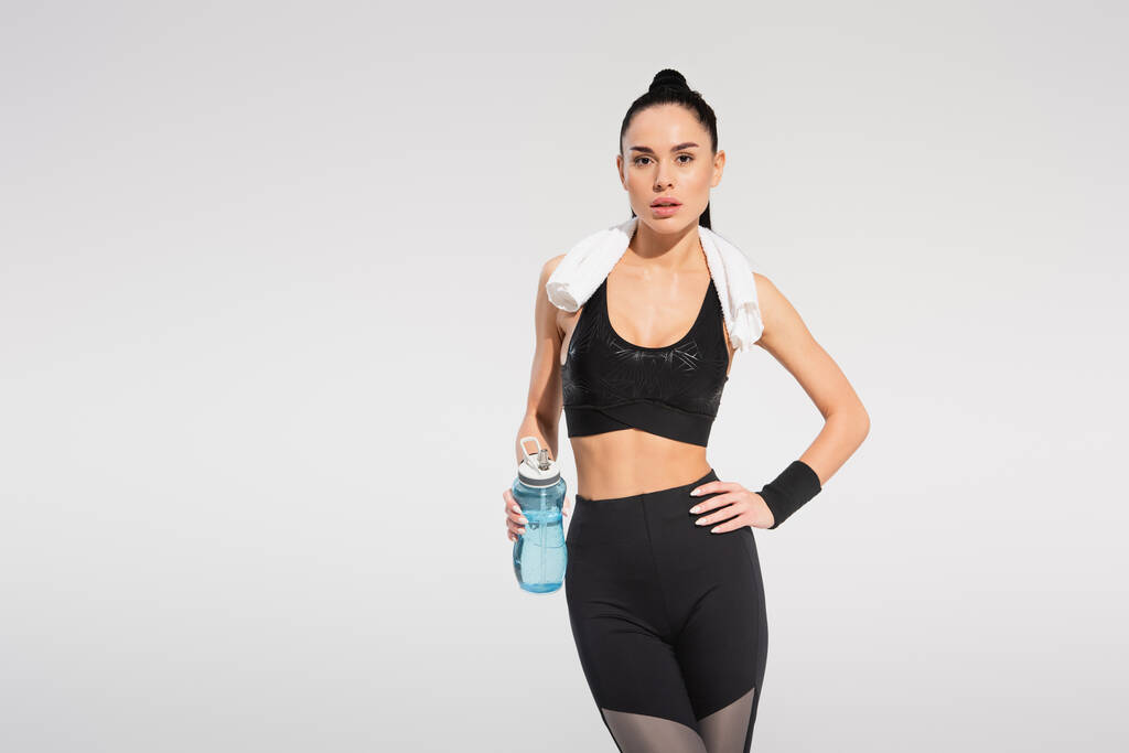 brunette sportswoman with towel on shoulders holding sports bottle while posing with hand on hip isolated on grey - Photo, Image