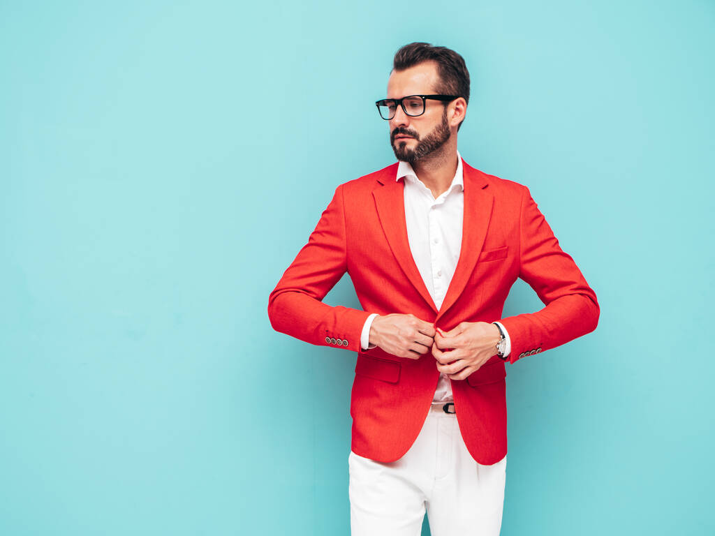 Portret przystojnego, pewnego siebie, modela lamberseksualnego hipstera.Ekskluzywny, nowoczesny mężczyzna ubrany w elegancki czerwony garnitur. Moda mężczyzna pozowanie w studio w pobliżu niebieskiej ściany w okularach przeciwsłonecznych - Zdjęcie, obraz