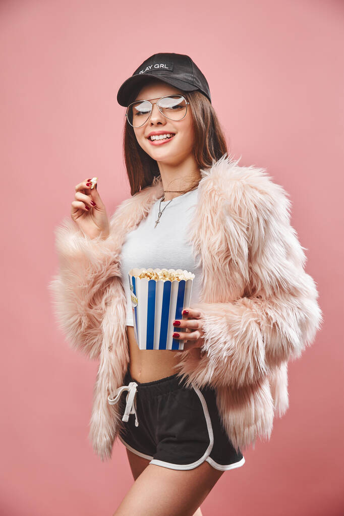 Aantrekkelijk brunette meisje in nep roze vacht met popcorn in de hand. Draag in zwarte korte broek witte top zwarte kap en bril. Ik kijk naar de camera. Studio roze achtergrond - Foto, afbeelding