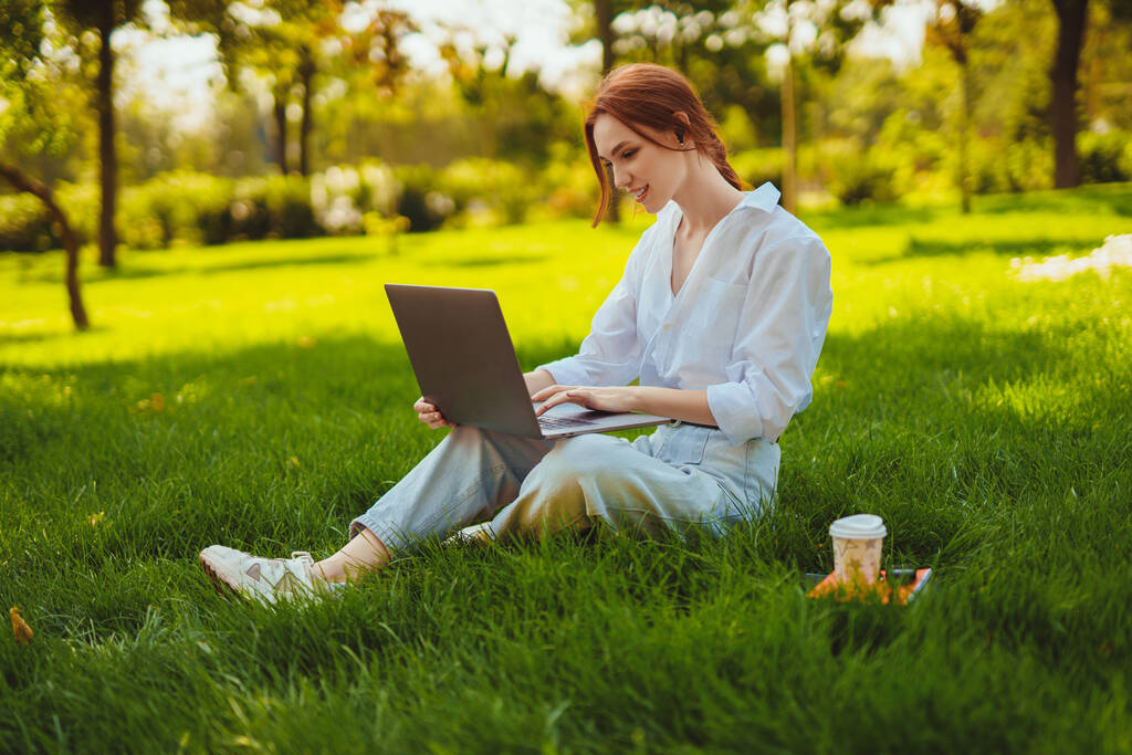 Красивая молодая красивая рыжая женщина в парке на открытом воздухе, используя ноутбук для изучения или работы в Интернете с помощью беспроводных наушников - Фото, изображение
