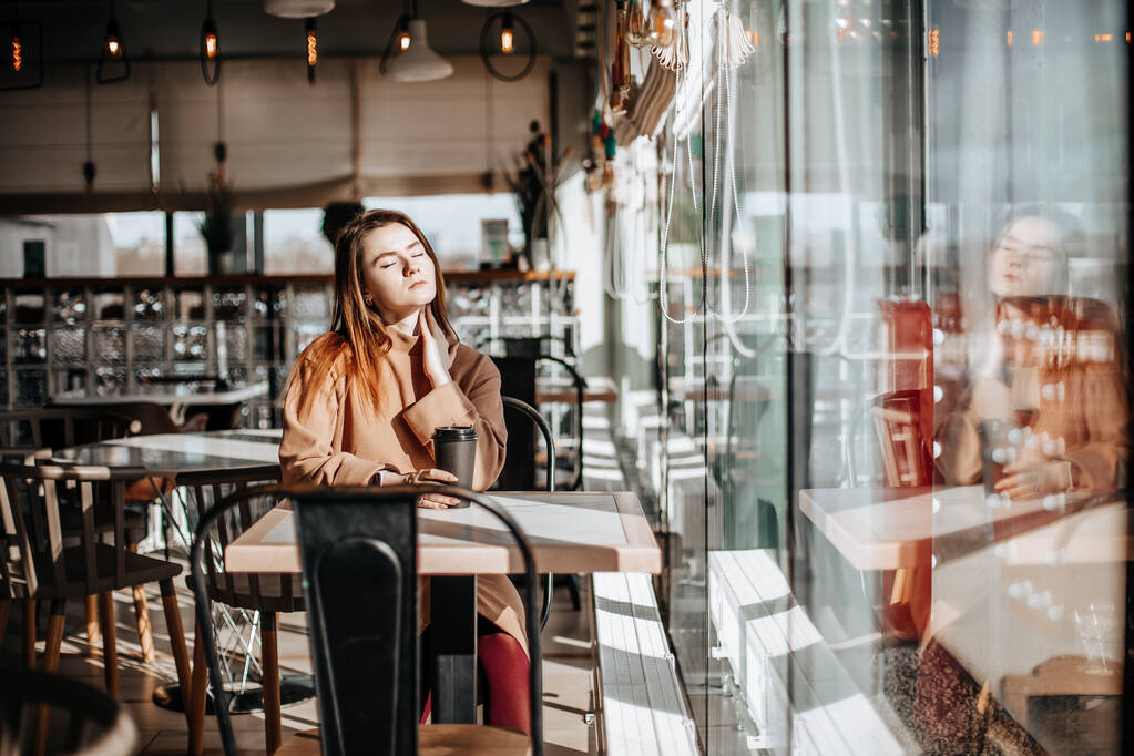 Stilvolles Mädchen sitzt in einem Café am Tisch und trinkt Kaffee. Coffee to go in einer Pappbecher. Frau mit roten Haaren im beigen warmen Anzug in gemütlicher Atmosphäre. Moderner Innenraum. Ruhiger und angenehmer Zeitvertreib. - Foto, Bild