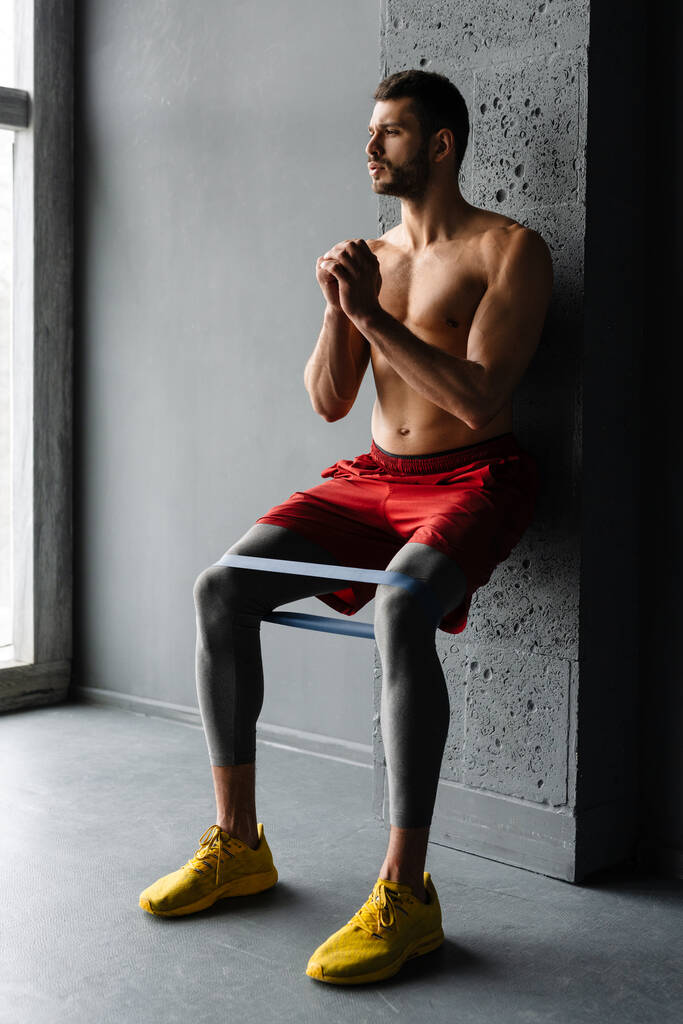 Jovem desportista sem camisa fazendo exercício com expansor fita elástica dentro de casa - Foto, Imagem
