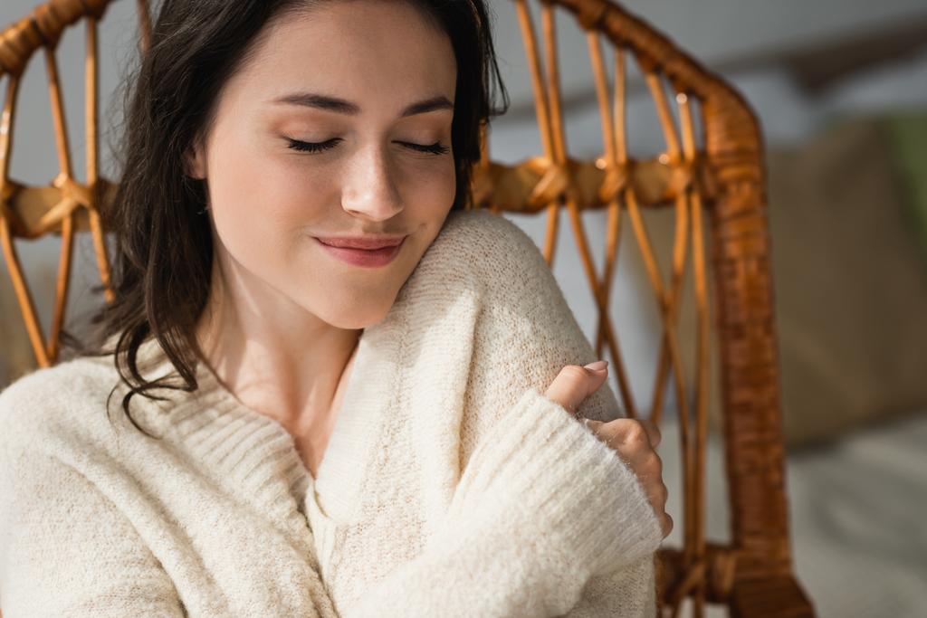 potěšená žena v teplém svetru objímající se při odpočinku doma se zavřenýma očima - Fotografie, Obrázek