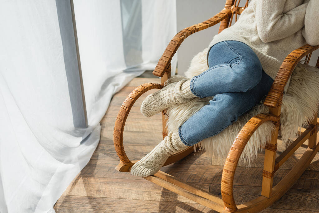 vista ritagliata di donna in jeans e calzini caldi seduti sulla sedia a dondolo - Foto, immagini
