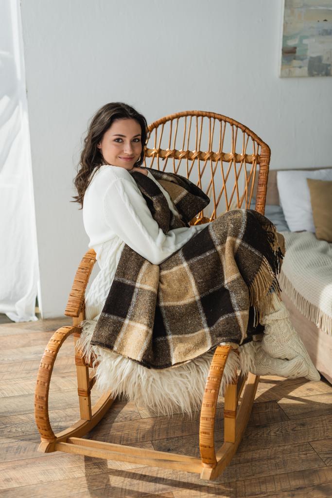 pleased woman looking at camera while sitting in wicker chair under plaid blanket - Photo, Image