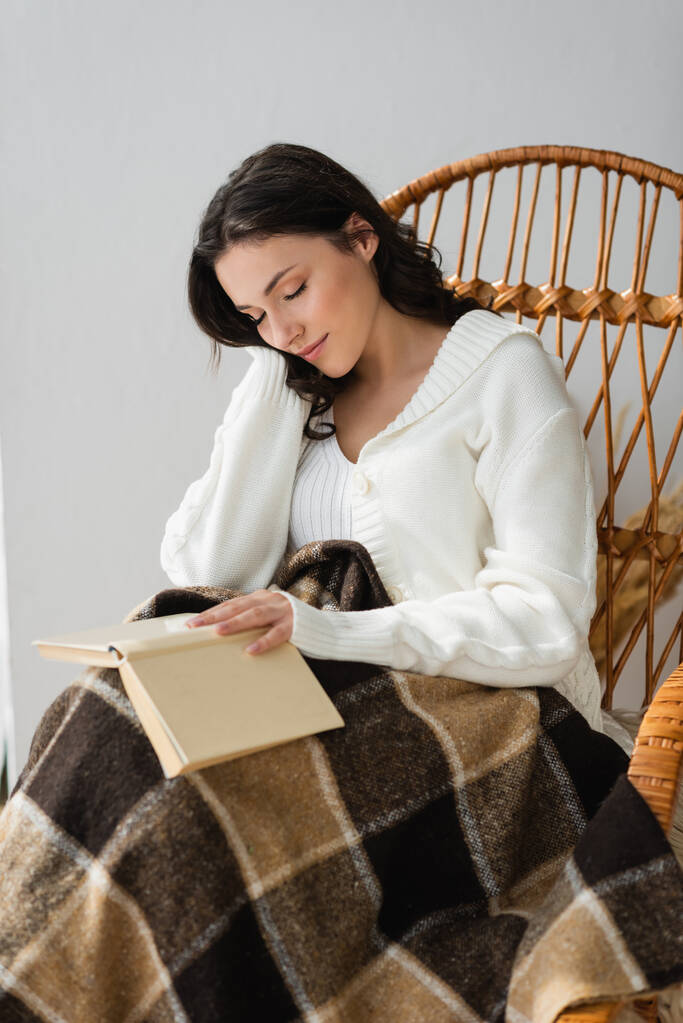 junge Frau mit Buch schläft im Korbstuhl unter karierter Decke - Foto, Bild