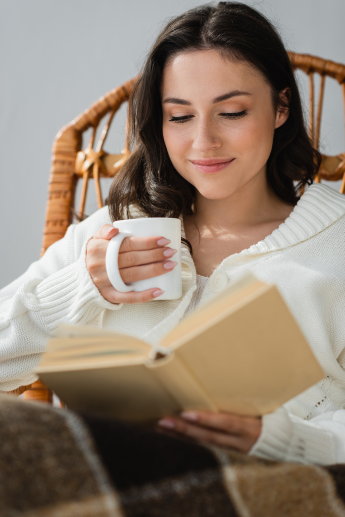 счастливая женщина с чашкой горячего напитка, читающая размытую книгу дома - Фото, изображение