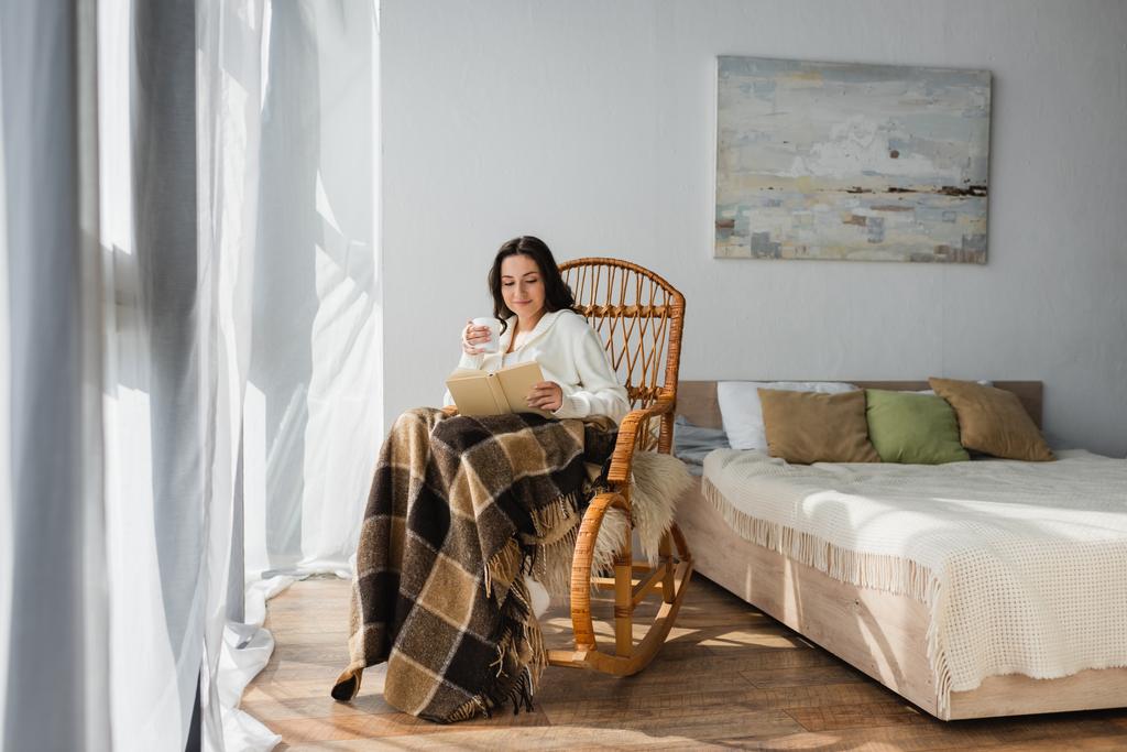 mujer morena leyendo libro en el dormitorio mientras está sentado en mecedora bajo manta a cuadros - Foto, Imagen