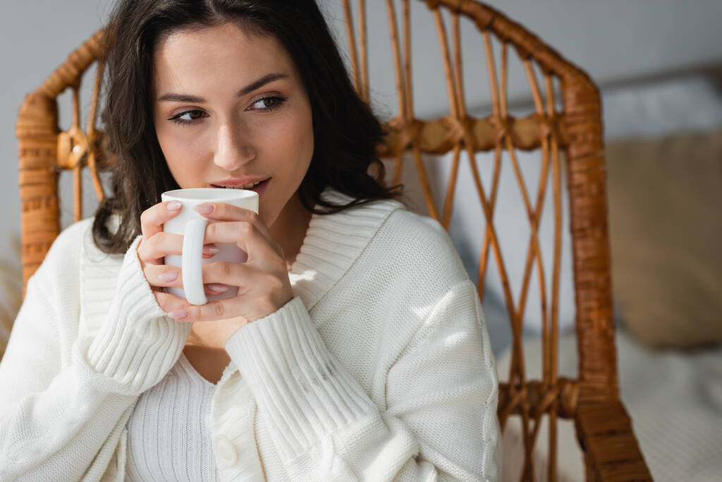 ブルネット女性でホワイトカーディガンで座っているウィッカーチェアと飲み物暖かいお茶 - 写真・画像