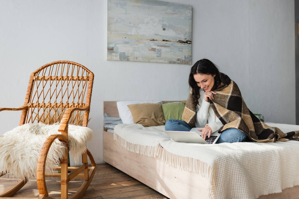 cheerful woman using laptop on bed near wicker chair  - Photo, Image