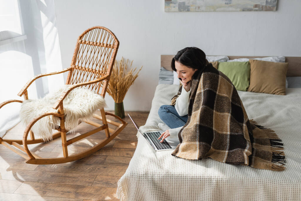 uśmiechnięta kobieta siedzi na łóżku przy bujanym krześle i używa laptopa - Zdjęcie, obraz