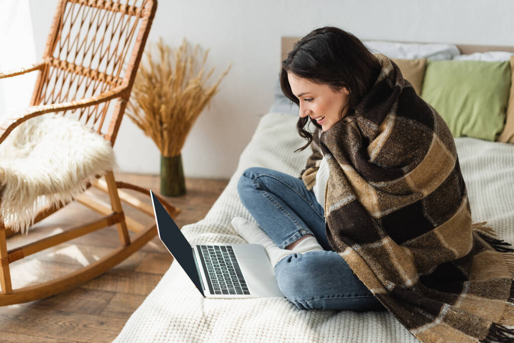 glimlachende vrouw op zoek naar laptop met leeg scherm terwijl zitten op bed onder geruite deken - Foto, afbeelding