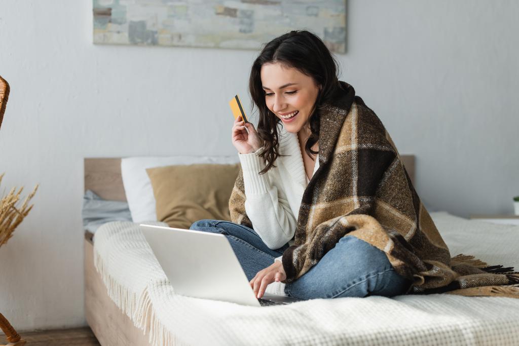 potěšená žena sedí na posteli pod teplou přikrývkou, zatímco drží kreditní kartu v blízkosti notebooku - Fotografie, Obrázek