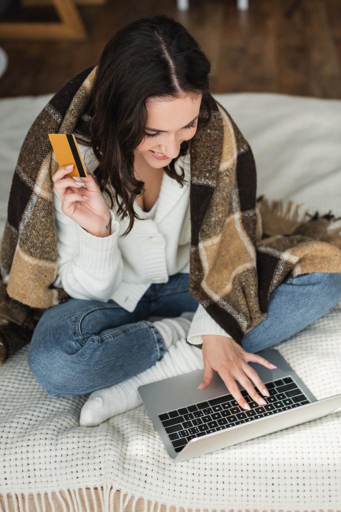 счастливая женщина держит кредитную карту и печатает на ноутбуке на кровати под клетчатым одеялом - Фото, изображение