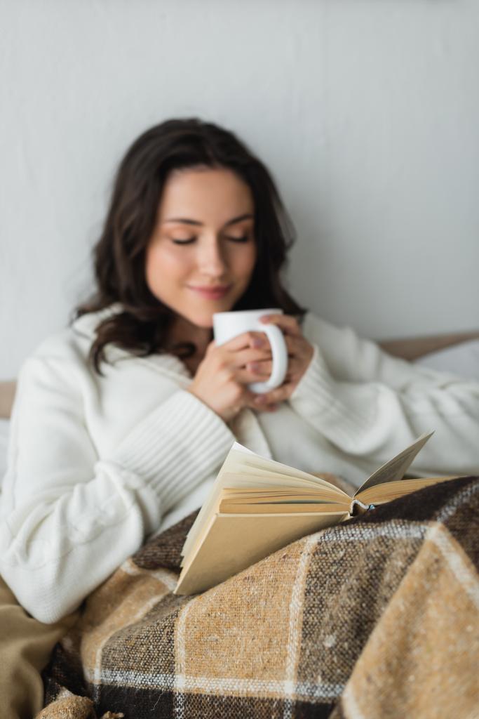 mulher desfocada com olhos fechados segurando xícara de chá enquanto descansa na cama com livro - Foto, Imagem