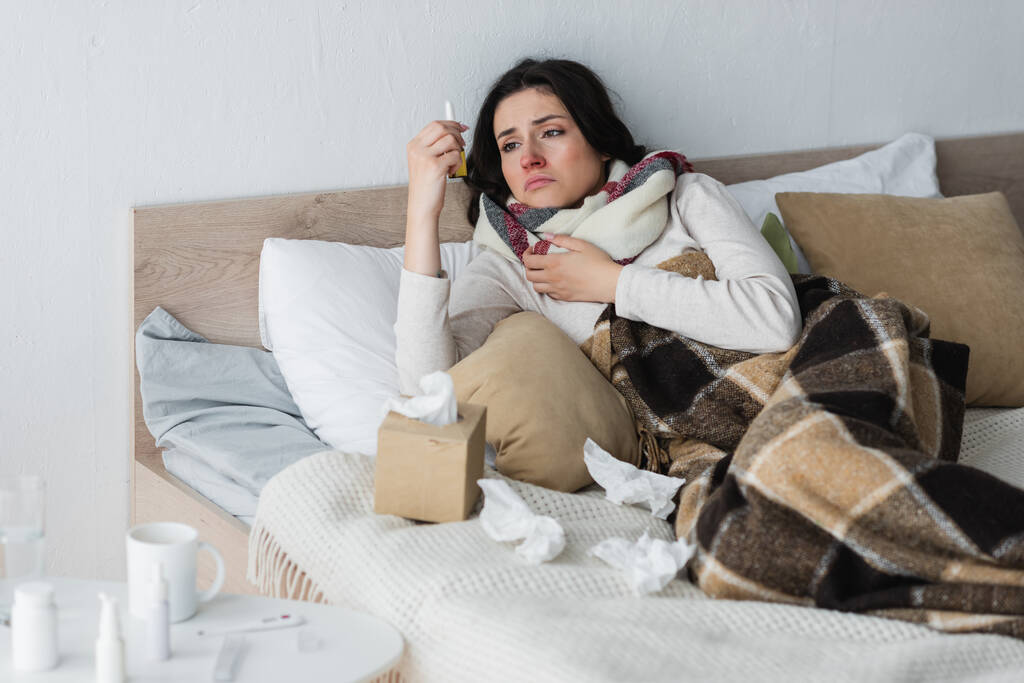 overstuur vrouw kijken naar neusspray terwijl liggend in bed in de buurt van verfrommeld papier servetten - Foto, afbeelding