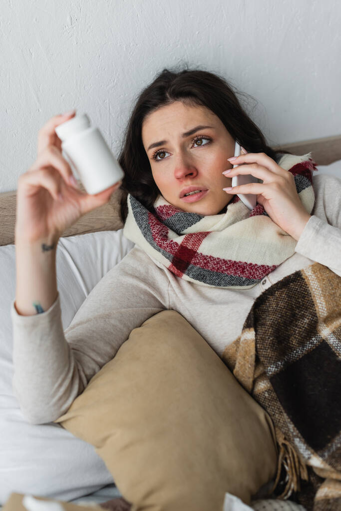 больная женщина с таблетками контейнер разговаривает по мобильному телефону, находясь под клетчатым одеялом - Фото, изображение