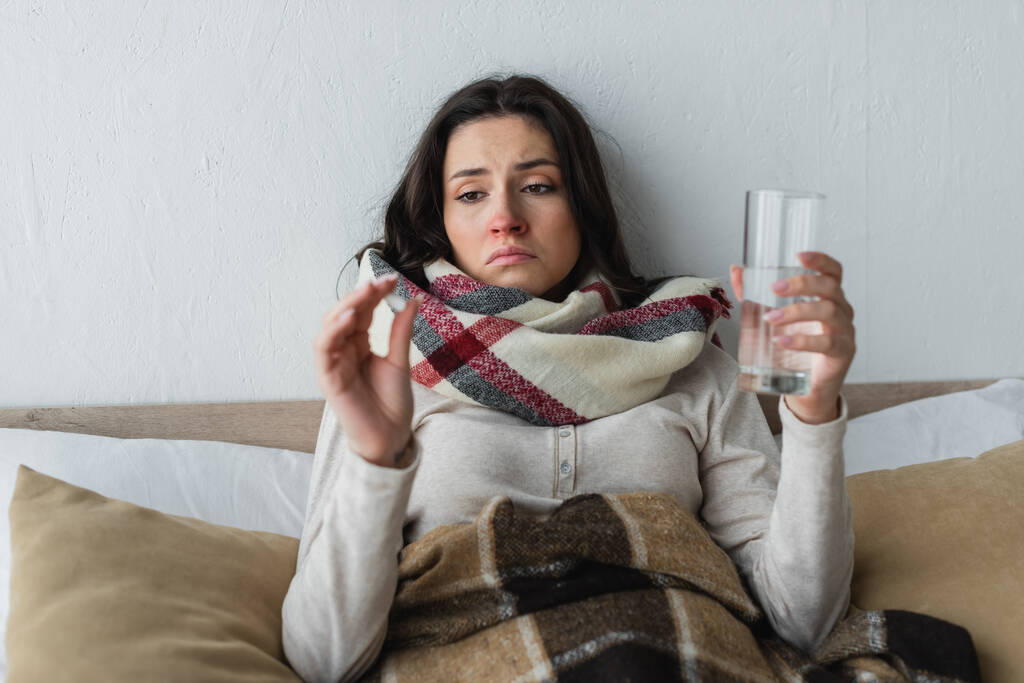 nemocná a smutná žena drží sklenici vody a pilulky, zatímco leží pod kostkovanou přikrývkou - Fotografie, Obrázek