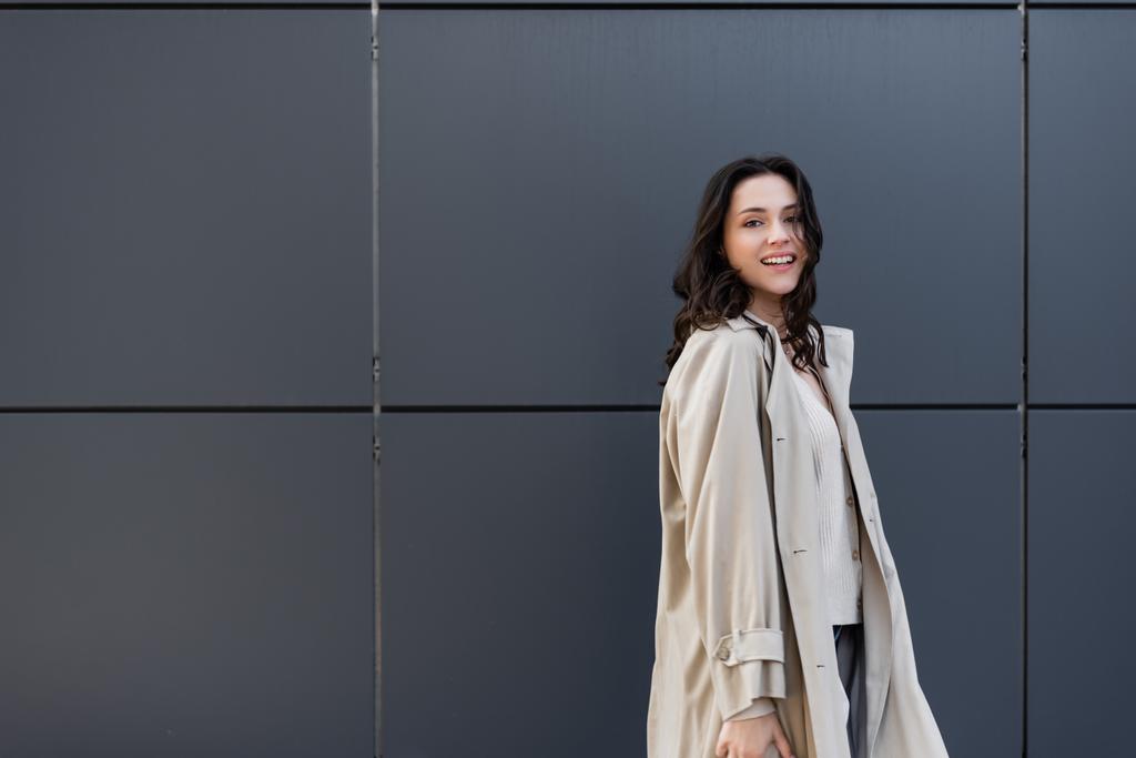 wesoła kobieta uśmiecha się do kamery podczas pozowania w beżowym płaszczu przy szarej ścianie - Zdjęcie, obraz