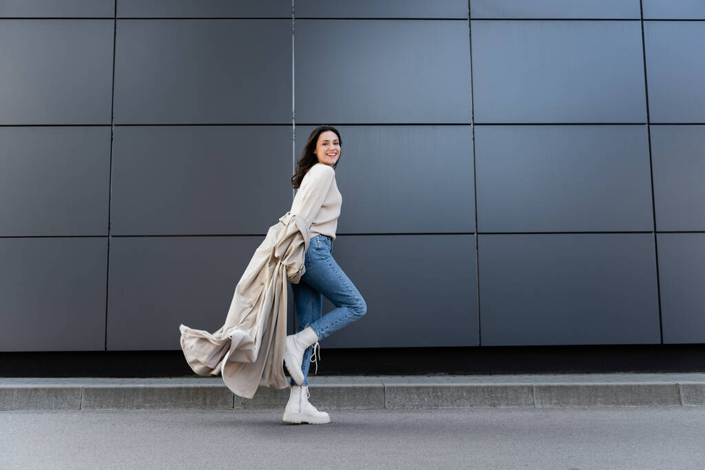 gelukkig vrouw in jeans en witte laarzen glimlachen op camera terwijl lopen langs grijze muur - Foto, afbeelding