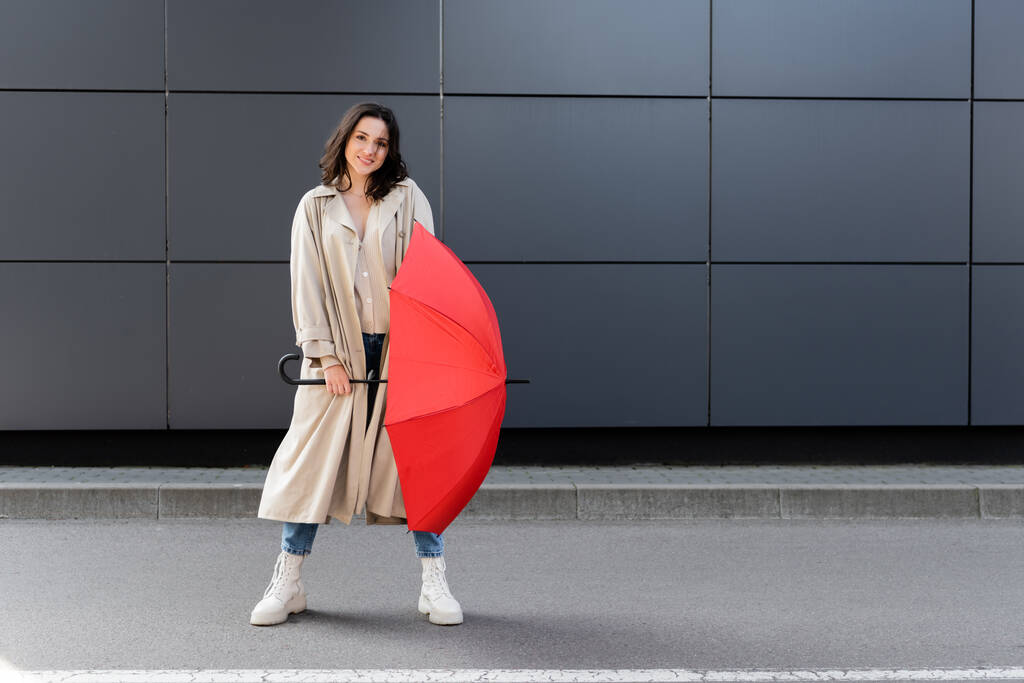 trendy woman in long coat and white boots posing with red umbrella near grey wall - Photo, Image