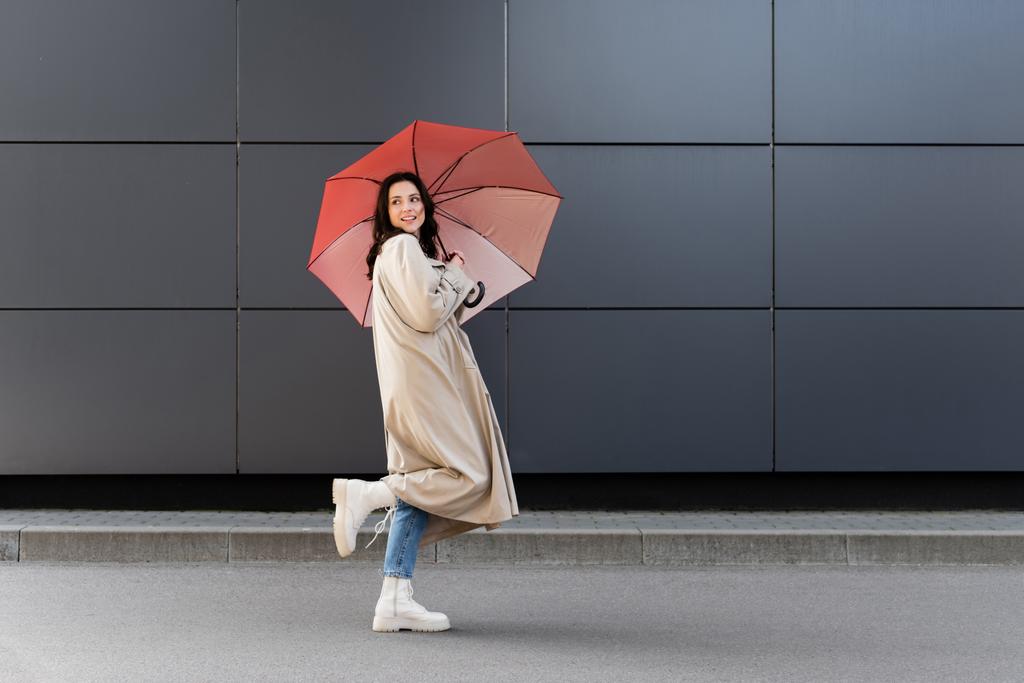 morena mujer en botas blancas y abrigo beige mirando hacia atrás bajo paraguas rojo - Foto, imagen