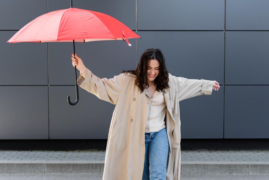 junge Frau in beigem Regenmantel lächelt, während sie mit rotem Regenschirm in der Nähe der grauen Wand posiert - Foto, Bild