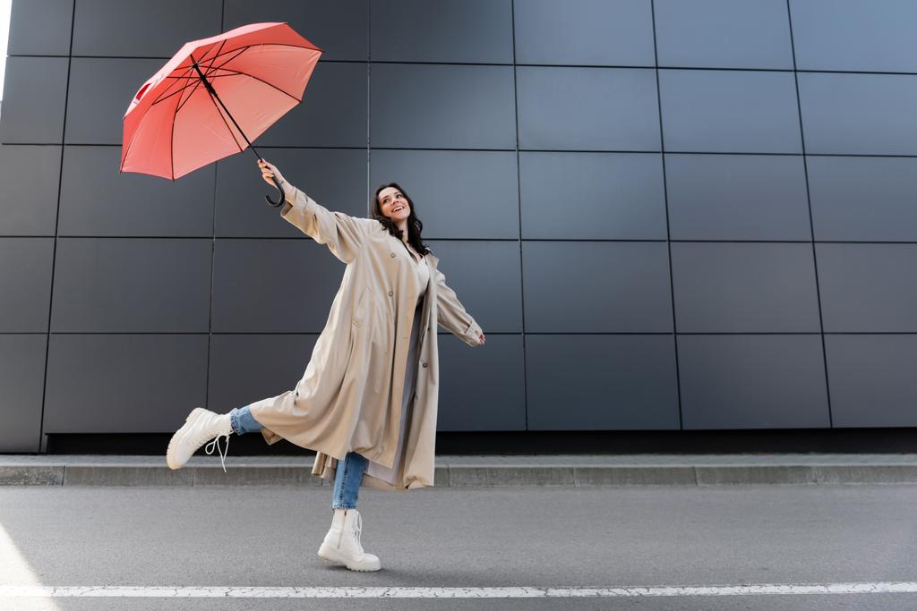 happy woman in beige coat and white boots posing with red umbrella in raised hand - Photo, Image