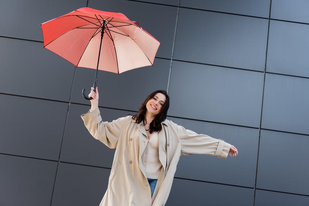 Brünette Frau im beigen Mantel lächelt in die Kamera unter rotem Regenschirm neben grauer Wand - Foto, Bild