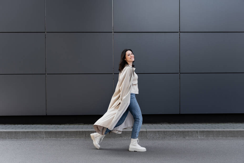 fröhliche Frau, die in langem Mantel und weißen Lederstiefeln an der grauen Wand entlang läuft - Foto, Bild
