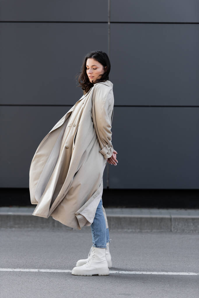 mulher na moda em longo casaco de pé com os olhos fechados e as mãos atrás de volta perto da parede cinza - Foto, Imagem