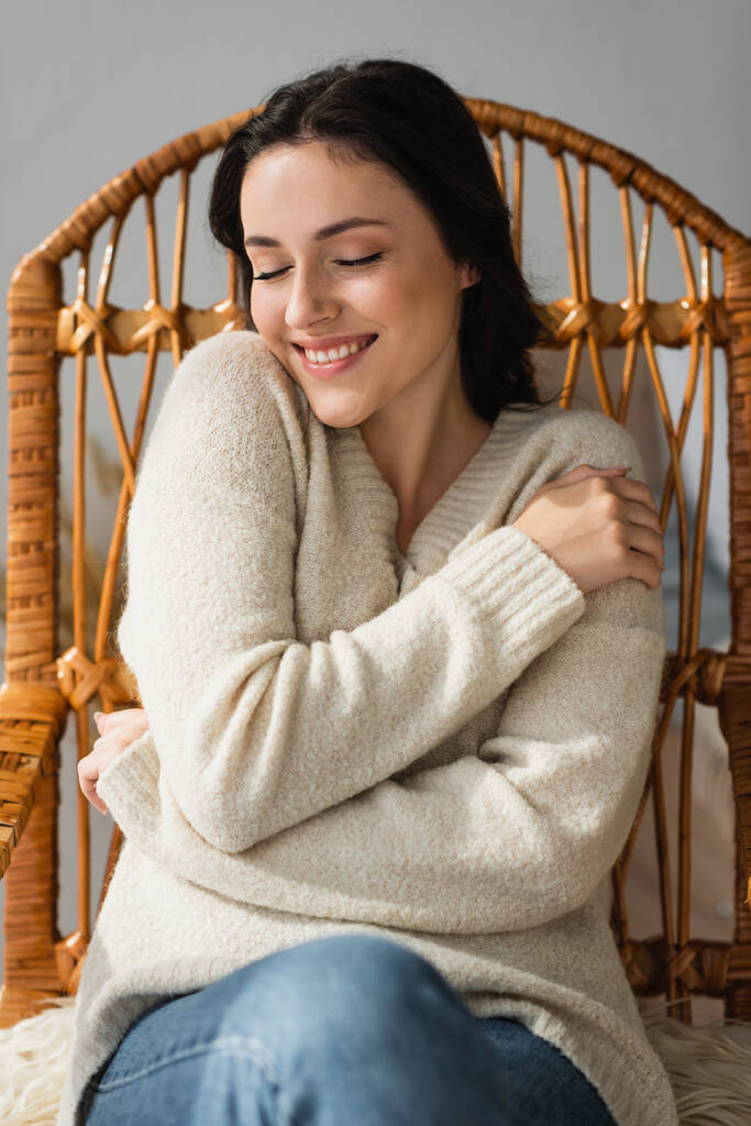 Улыбающаяся женщина в уютном свитере обнимается в плетеном кресле - Фото, изображение