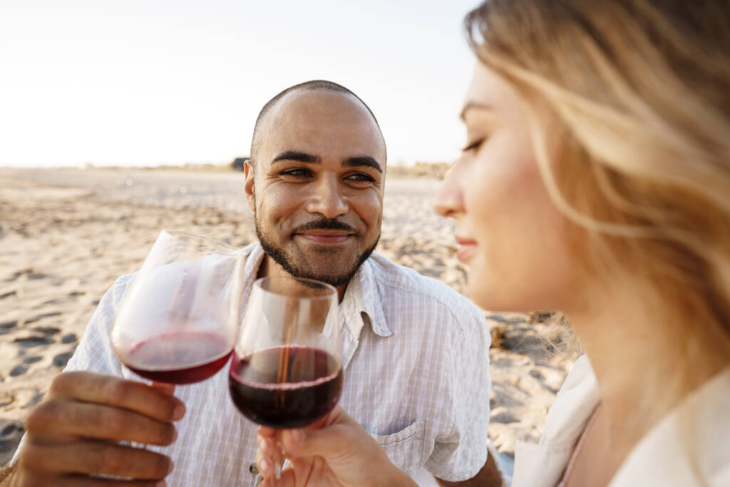 Portrait d'un jeune couple assis sur la plage et buvant du vin - Photo, image