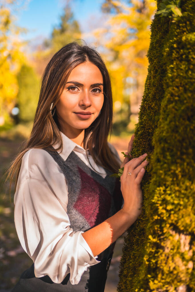 Yaşam tarzı, genç bir esmer üniversiteli kız, bir sonbahar öğleden sonra bir ağacın yanındaki parkta şık bir pozda. - Fotoğraf, Görsel