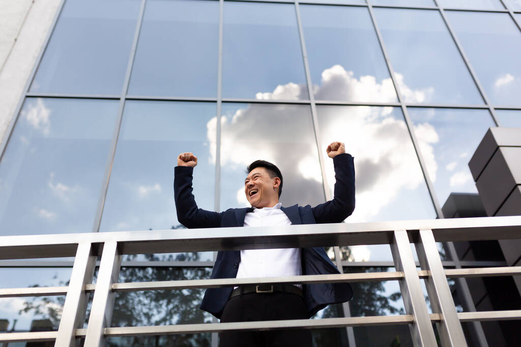 アジアのビジネスマンの上司は暗い色のビジネススーツでバルコニーの彼のオフィスセンターの近くに立つ彼の手を保持して勝利を祝う - 写真・画像