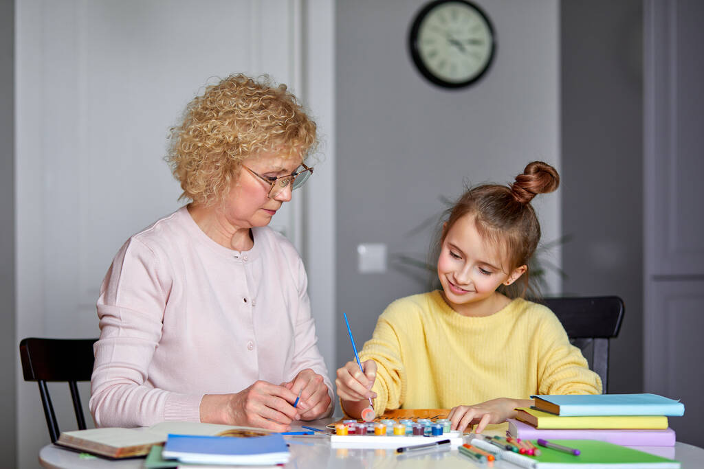 Красивая внучка и старшая бабушка сидят дома, рисуют, делают домашнюю работу - Фото, изображение