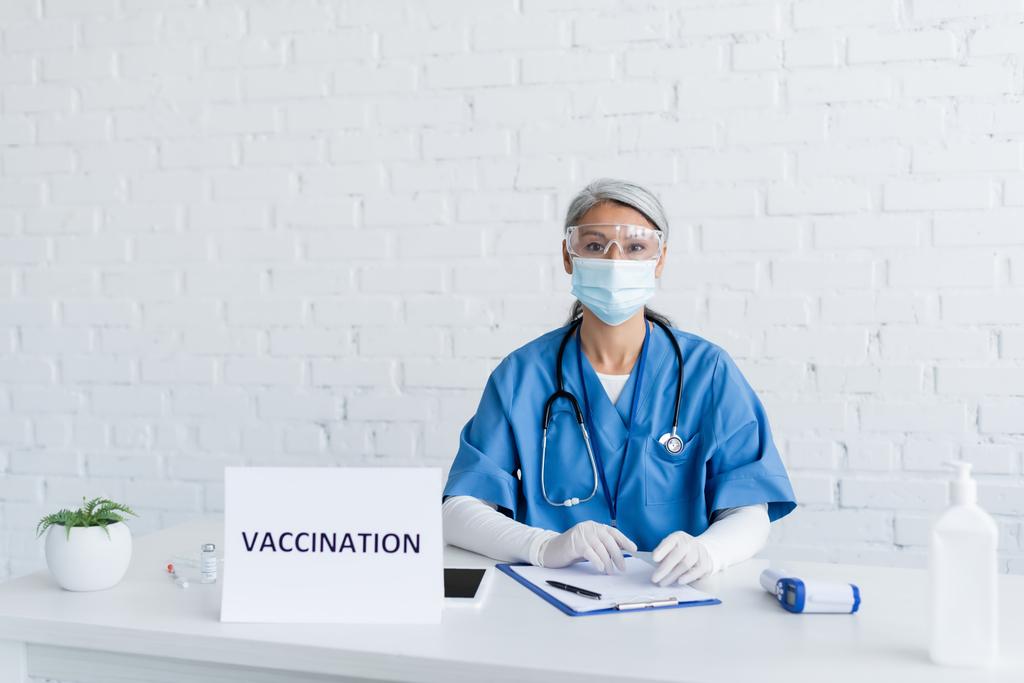 medico asiatico di mezza età in occhiali e maschera medica vicino targa segno con iscrizione di vaccinazione su scrivania - Foto, immagini
