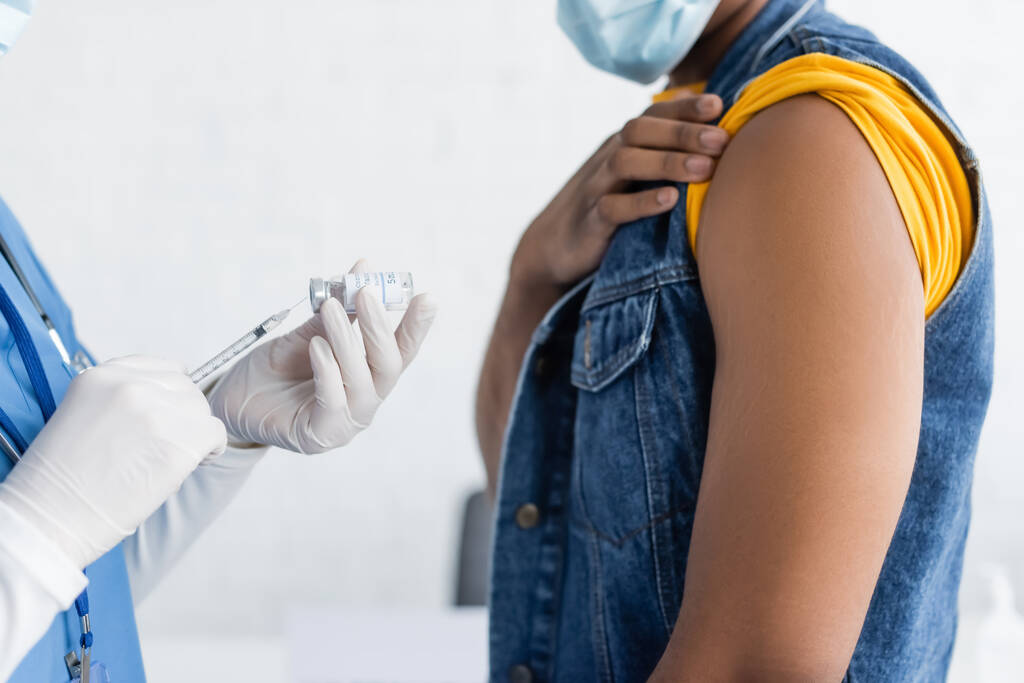 アフリカ系アメリカ人男性の近くにワクチンを保持するラテックス手袋の看護師のクロップドビュー医療マスク - 写真・画像