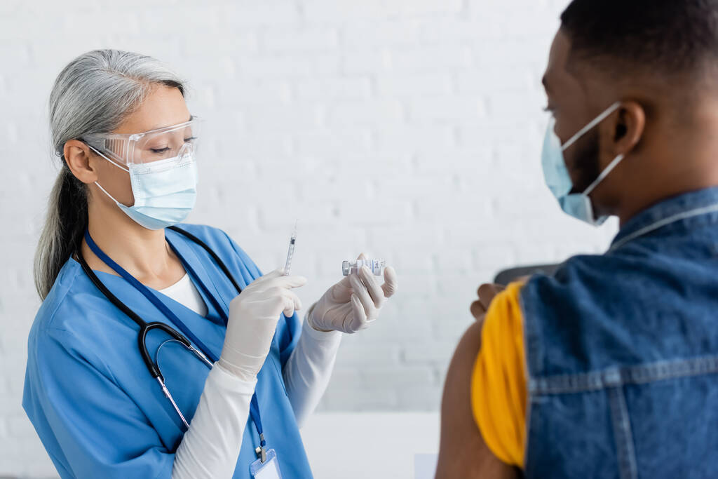asiático enfermera en médico máscara celebración jeringa y tarro con vacuna cerca borrosa africano americano hombre - Foto, imagen