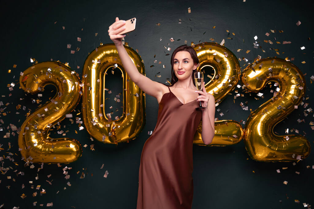 Hermosa mujer en vestido moderno de lujo haciendo selfie con champán delante de globos dorados 2022 aislados sobre fondo oscuro, confeti. - Foto, imagen