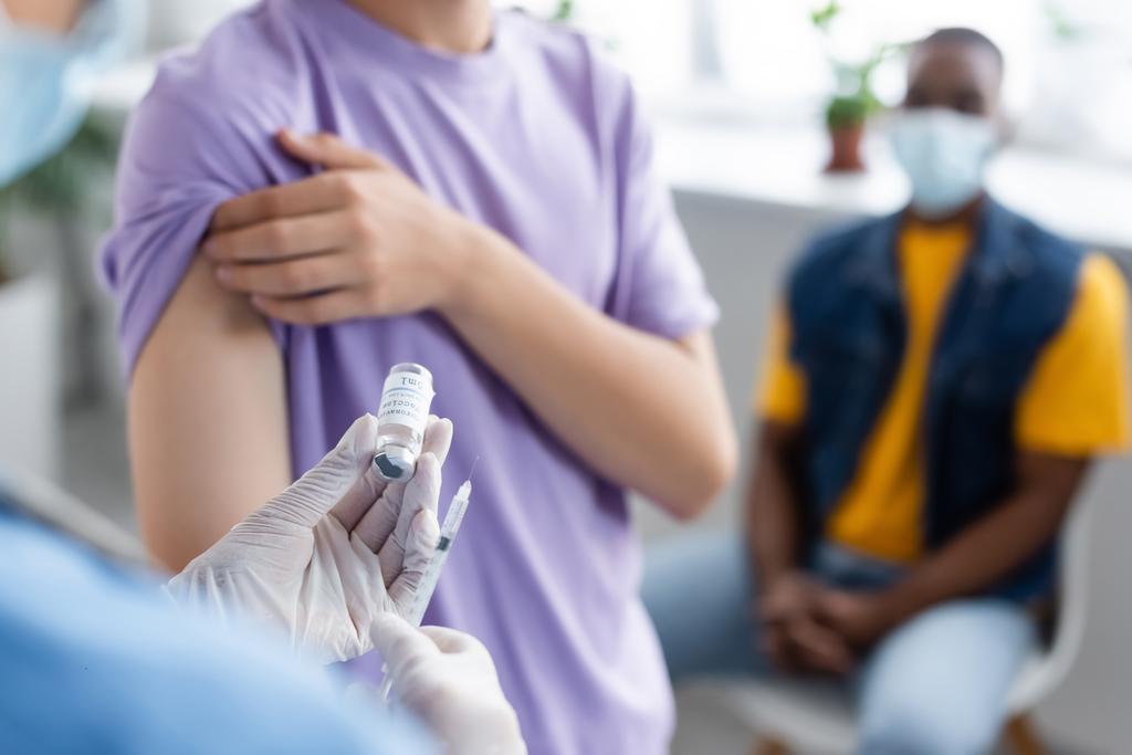 若い患者とアフリカ系アメリカ人男性の近くにワクチンを持っているラテックス手袋のぼやけた看護師 - 写真・画像