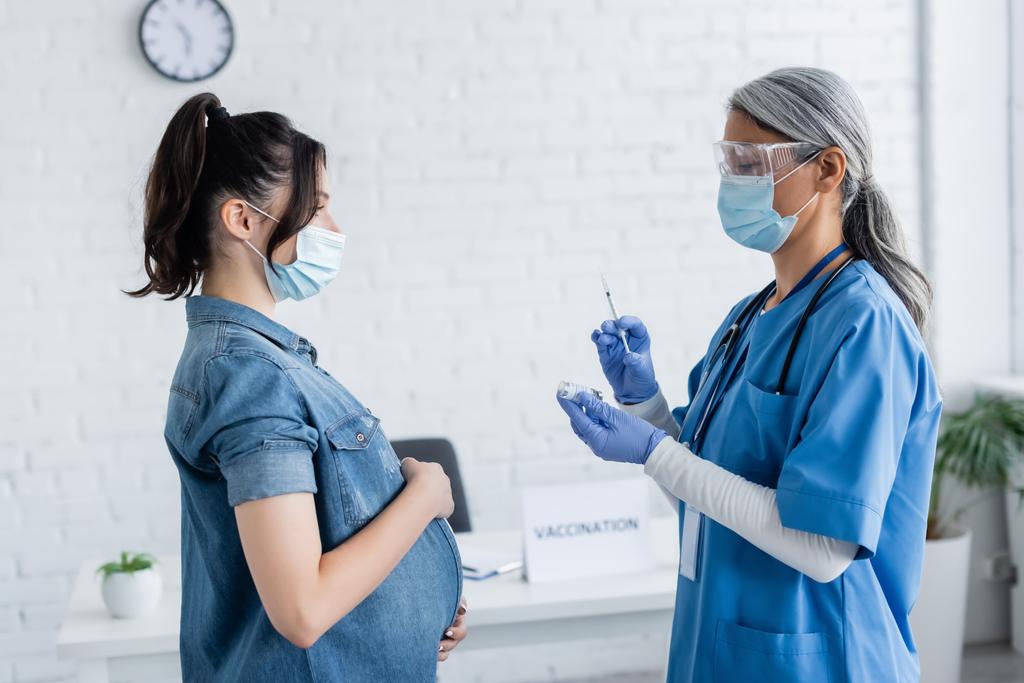 πλαϊνή όψη ώριμης νοσοκόμας με ιατρική μάσκα που κρατάει σύριγγα και βάζο με εμβόλιο κοντά σε έγκυο γυναίκα - Φωτογραφία, εικόνα