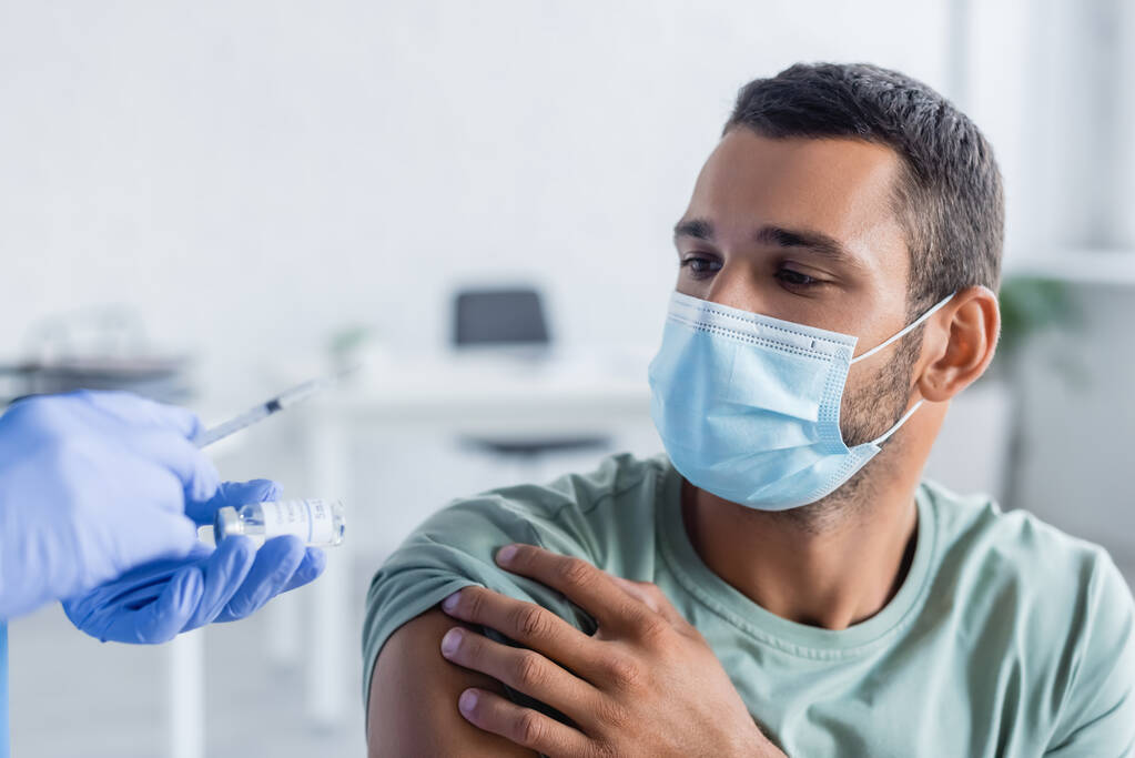 orvos kezében fecskendő és üveg oltóanyag közelében fiatal férfi orvosi maszkban - Fotó, kép