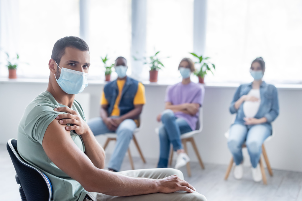 νεαρός άνδρας με προστατευτική μάσκα κοιτάζοντας την κάμερα κοντά σε θολή πολυεθνικά άτομα που κάθονται στην κλινική, έννοια του εμβολιασμού - Φωτογραφία, εικόνα
