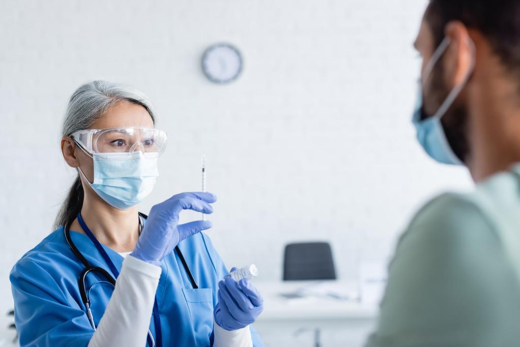 mature asiatique médecin dans médical masque et lunettes préparation seringue avec vaccin près flou homme - Photo, image